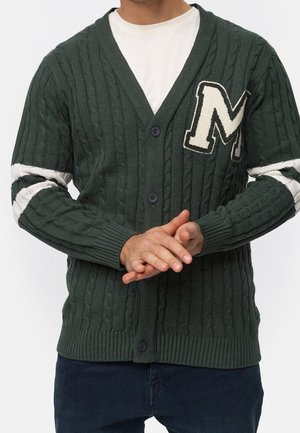 Mężczyzna w ciemnozielonym kardiganie z grubym splotem, ozdobionym dużą białą naszywką w kształcie litery "M" oraz białymi paskami na rękawach, pod którym ma na sobie białą koszulę.