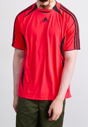 Mies, jolla on päällään punainen Adidas-urheilupaita, jossa mustat raidat olkapäillä, ja oliivinvihreät housut, seisoo yksivärisen taustan edessä.