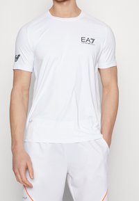 Vit sporttröja i ett strukturerat, ventilerande tyg. Har korta ärmar och EA7-logotypen tryckt på vänster sida.