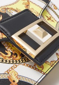 Zbliżenie na zapięcie torebki w kolorach czarnym, białym i złotym z kwadratową, pozłacaną klamrą oraz skórzanym tłem z wzorem w stylu barokowym.