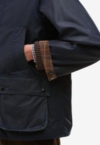 Ναυτικό αδιάβροχο μπουφάν με λεπτομέρεια καρό στα μανίκια. Διαθέτει μεγάλη μπροστινή τσέπη και κουμπώματα με πίεση. Λείο ύφασμα με χαλαρή εφαρμογή.