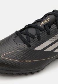 Černé fotbalové kopačky F50 vyrobené z materiálu Fiberskin, s texturovaným povrchem, černými tkaničkami a stříbrnými pruhy se zlatým akcentem.