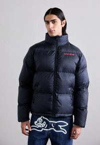 Doudoune navy avec col montant et fermeture éclair. Présente des sections duveteuses, deux poches latérales et un texte rouge "glace" sur la poitrine.