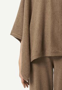 Person trägt locker sitzendes, hellbraunes Strickoberteil und Hose, der linke Arm entspannt und sichtbar vor einem einfarbigen beigen Hintergrund.