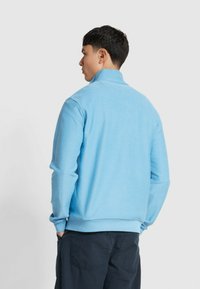 Lichtblauwe zip-up sweatshirt met een subtiele textuur, hoge kraag en geribde boorden, gecombineerd met donkere broeken. Achteraanzicht weergegeven.