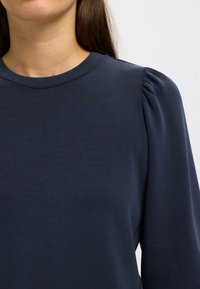 Granatowy top z długim rękawem o gładkiej teksturze, okrągłym dekolcie i delikatnie plisowanych detalach na ramionach. Prosty, minimalistyczny wzór.