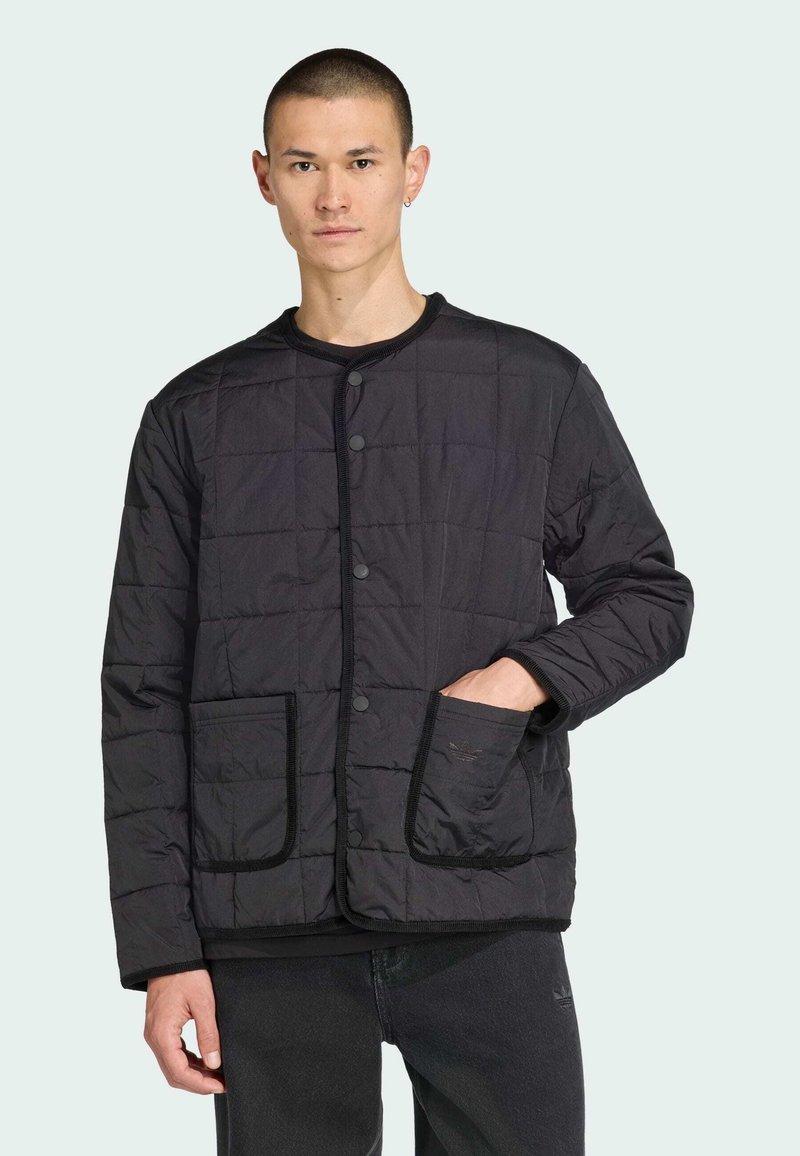 Le modèle porte une veste matelassée noire avec des boutons et des poches, se tenant devant un fond vert clair, dégageant un look décontracté et élégant.