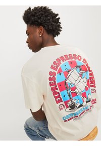 Cremefarvet t-shirt med et stort grafisk motiv på ryggen. Designet inkluderer en kaffekande og kop, med røde og blå accenter mod en ternet baggrund.
