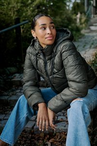 Pufferjakke i mørk olivengrøn med høj krave, lynlåslukning foran og ribbede manchetter. Bæret med lyseblå slidte jeans.