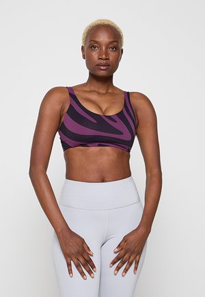 Atletická žena s krátkými blond vlasy, která má na sobě fialovo-černý sportovní podprsenku se vzorem a světle šedé legíny s vysokým pasem, stojící před jednobarevným pozadím.