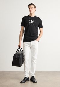 Černé bavlněné tričko s grafikou pavouka, bílé džíny, černé kožené boty a černá kožená cestovní taška. Jednoduchý design, moderní střih.