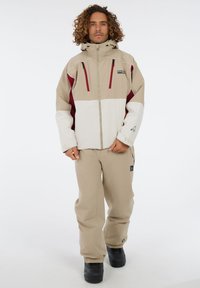 Beige en witte waterdichte jas met rode accenten, met ritszakken en een capuchon; gecombineerd met bijpassende beige broek en zwarte laarzen.