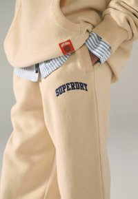 Béžová flísová mikina s rebrovanými manžetami, navrstvená cez modrú pruhovanú košeľu. Tepláky sú s námorníckym logom "SUPERDRY" na stehne.