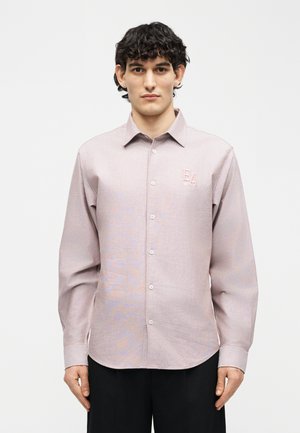 Göndör hajú férfi világos rózsaszín gombos ingben, amelynek mellkasán "EA" logó látható, egyszínű fehér háttér előtt áll.