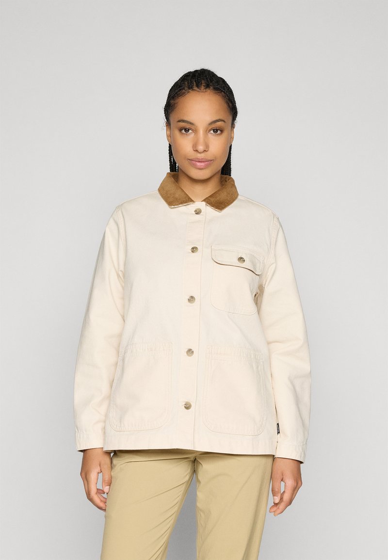 Vrouw in een crème kleurige jas met een bruine corduroy kraag, met een knoopsluiting aan de voorkant, en beige broek, staande tegen een effen achtergrond.