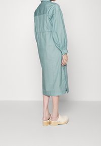 Donna in un vestito midi a maniche lunghe di un delicato blu chiaro con sottili righe verticali e zoccoli in legno beige, in piedi contro uno sfondo bianco semplice.