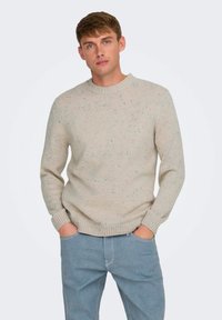 Suéter de punto beige con cuello redondo y puños acanalados. Cuenta con motas multicolores por todo el tejido. Combinado con pantalones de denim azul claro.