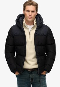 Czarna kurtka puchowa z zapięciem na zamek, odpinanym kapturem i kieszeniami na piersi. Nosi na sobie kremowy sweter z ćwierć zamkiem. Dżinsy zapinane na guziki i zegarek na ręku.