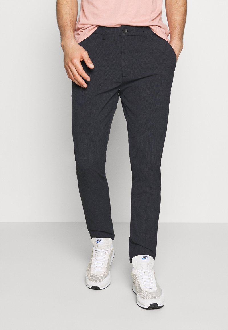 Donkergrijze, slim-fit broek met een gestructureerde stof, voorzien van een knoopsluiting aan de voorkant en zijzakken, gecombineerd met witte sportschoenen.