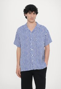 Kék és fehér hullámos mintás rövid ujjú ing, gallérral, elülső zsebbel, gombos záródással, könnyű anyagból, fekete nadrággal viselve.