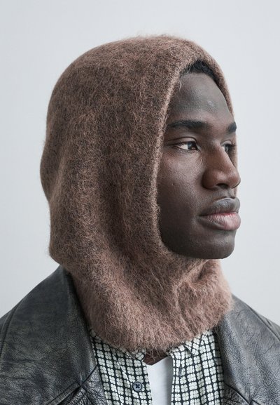 Mladý černoch v hnědé chlupaté kapuci, vzorované košili a černé kožené bundě, který zamyšleně hledí stranou na jednoduchém pozadí.