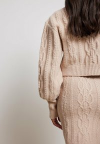 Pull tricoté beige clair avec motif torsadé, design court et manches bouffantes. Texture douce avec taille côtelée et détail au dos.