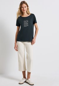T-shirt vert foncé à manches courtes avec un motif graphique blanc. Associé à un pantalon large crème et des baskets blanches, présentant un tissu texturé.