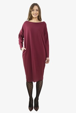 Bordowa, luźna sukienka z miękkiego materiału, z dekoltem w łódkę, długimi rękawami i bocznymi kieszeniami. Modelka ma na sobie dopasowane, czarne rajstopy w wzory.
