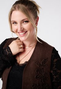 Glunderende vrouw met blond haar in een paardenstaart draagt gouden hoepel oorbellen, gelaagde gouden kettingen, een ring, een zwarte kanttop en een bruine vest met bloemenappliqué.