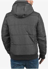 Pelēka polsterēta jaka ar kapuci, kas izgatavota no faktūrēta materiāla, ar ribotām aprocēm un pieguļošu dizainu. Redzams šuvums un kviltētas daļas.