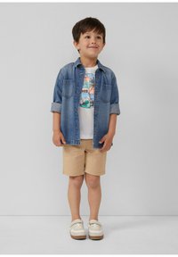 Jeanshemd mit hochgekrempelten Ärmeln über einem grafischen weißen T-Shirt, beigen Shorts und weißen Schuhen mit beigen Akzenten. Einfaches und lässiges Outfit.