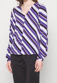 Long-sleeve top in purple, blue, and black stripes; V-neck design with a relaxed fit and smooth texture. Paired with black trousers.