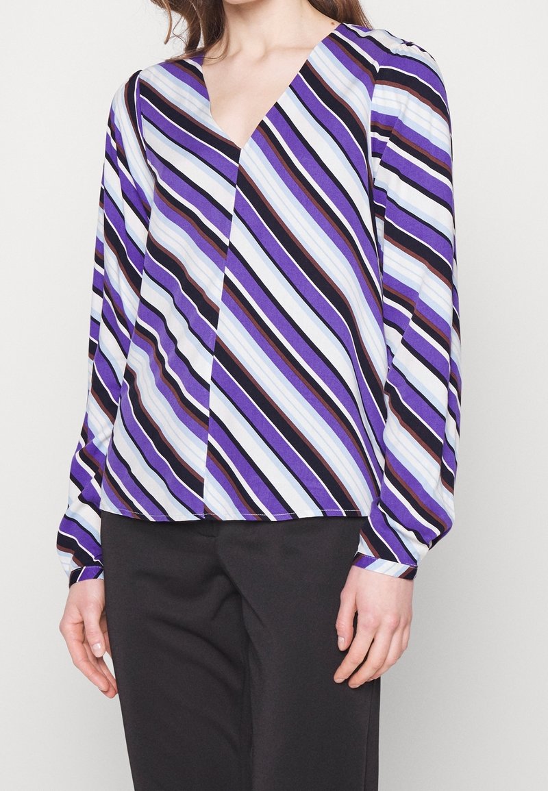 Long-sleeve top in purple, blue, and black stripes; V-neck design with a relaxed fit and smooth texture. Paired with black trousers.