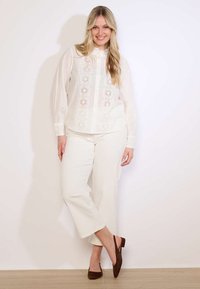 Usmievavá žena s dlhými blond vlasmi, oblečená v bielej blúzke z čipky, bielych krátkych nohaviciach a hnedých balerínach s otvorenou päty, stojaca pri bielej stene.