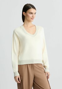 Maglione color crema a V con polsini e orlo a coste, caratterizzato da una texture morbida e una vestibilità rilassata, abbinato a pantaloni beige.