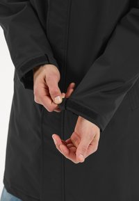 Svart vattentät jacka med en mjuk yta; närbild av händer som justerar kardborrefästen vid ärmsluten; slät textur och skräddarsydd passform.