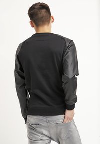 Sort sweatshirt med læderlange ærmer; ribbet halsudskæring og kant; tætsiddende design; båret med lysegrå jeans, set fra ryggen.