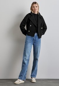 Svart kort dubbelknäppt jacka med silverknappar, matchad med ljusblå raka jeans och beige sneakers.