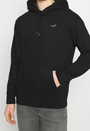 Hombre vistiendo una sudadera con capucha negra y un logo de ave blanca en el pecho, con la mano en el bolsillo delantero, acompañado de jeans grises, frente a un fondo claro y sencillo.