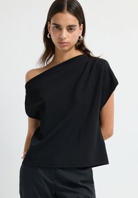 Donna con lunghi capelli scuri che indossa una maglietta nera senza spalle e pantaloni neri, accessoriate con orecchini a cerchio di argento, su uno sfondo bianco semplice.