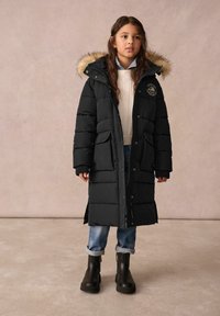 Jeune fille portant un long manteau matelassé noir avec capuche doublée de fourrure, pull crème, jean retroussé et bottes noires, debout à l'intérieur.