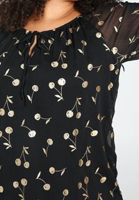 Blouse noire avec manches courtes transparentes, ornée d'un motif floral cerise doré. Encolure froncée avec un lien au centre devant.