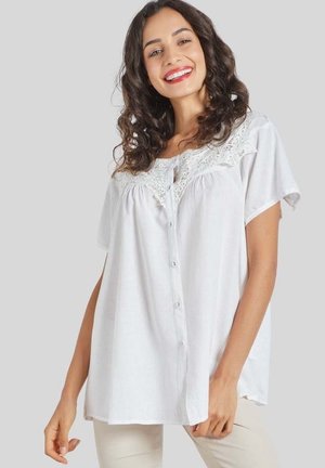 Mujer con cabello rizado y oscuro que lleva una blusa blanca suelta abotonada con detalles de encaje, sonriendo frente a un fondo liso gris claro.