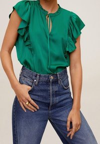 Blouse en satin vert avec manches à volants, décolleté en trou de serrure ; associée à un jean denim bleu taille haute. Texture lisse, couleur vibrante.