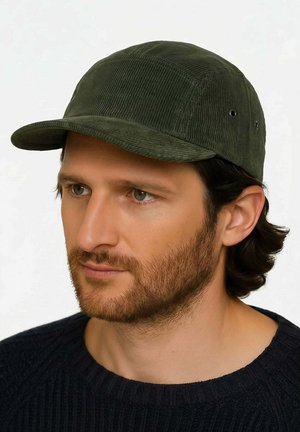 Man with light beard and medium-length hair wears dark green corduroy baseball cap and black knit sweater, facing slightly left.