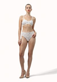 Donna in piedi che indossa lingerie in pizzo bianco con slip a vita alta e tacchi nudi contro uno sfondo chiaro e uniforme.