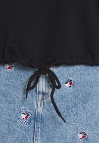 Czarny krótki top z ściągaczem u dołu, w połączeniu z jasnoniebieskimi szortami dżinsowymi z haftowanymi czerwonymi i granatowymi sercami oraz napisem "TOMMY JEANS".