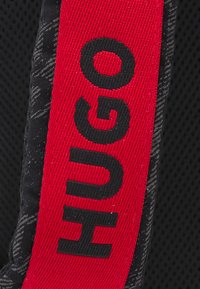 Tessuto nero con un motivo testurizzato, caratterizzato da una banda rossa e nera che mostra la parola "HUGO" in lettere in grassetto.