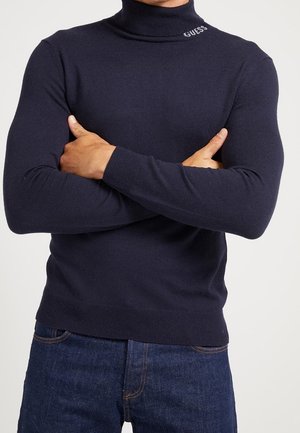 Mann trägt einen navyblauen Rollkragenpullover mit "GUESS"-Text am Kragen, die Arme über der Brust verschränkt, kombiniert mit dunkelblauen Jeans.