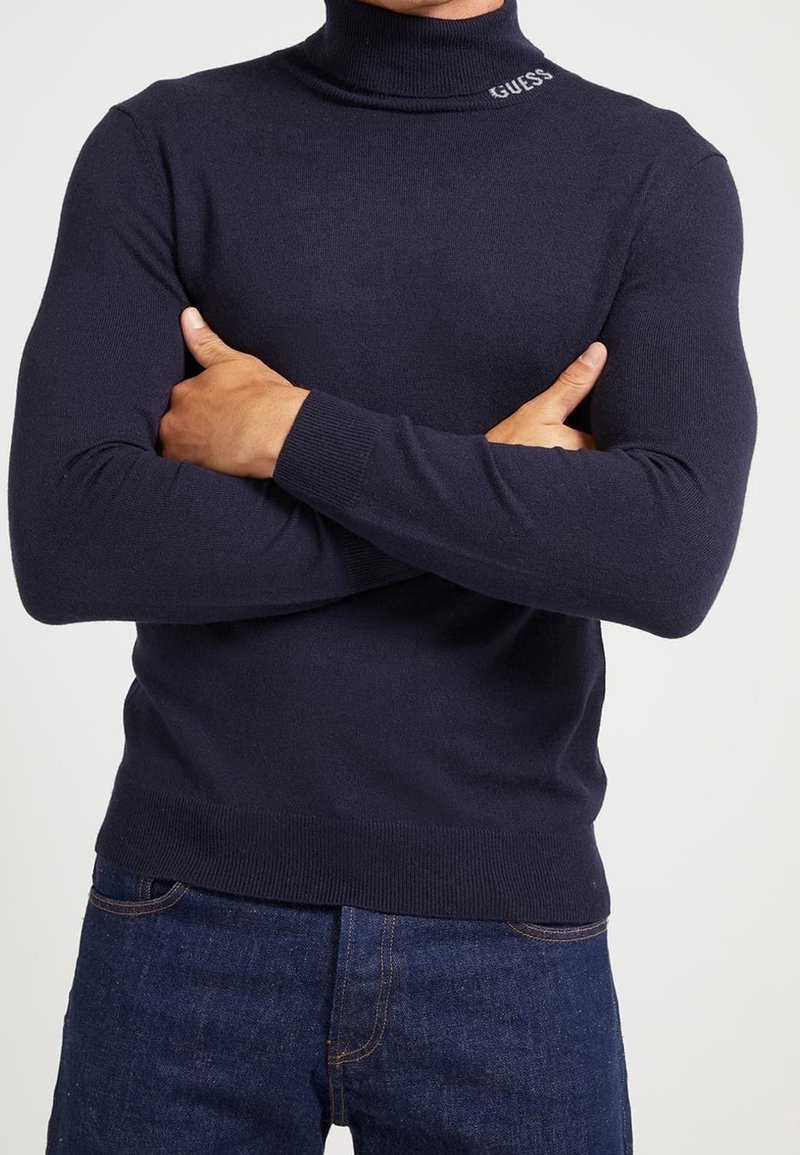 Uomo che indossa un dolcevita blu navy con il testo "GUESS" sul colletto, braccia incrociate sul petto, abbinato a jeans blu scuro.